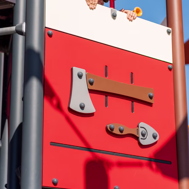 Child hidden behind red and white playground wall, peeking through toy binoculars, under clear blue sky.