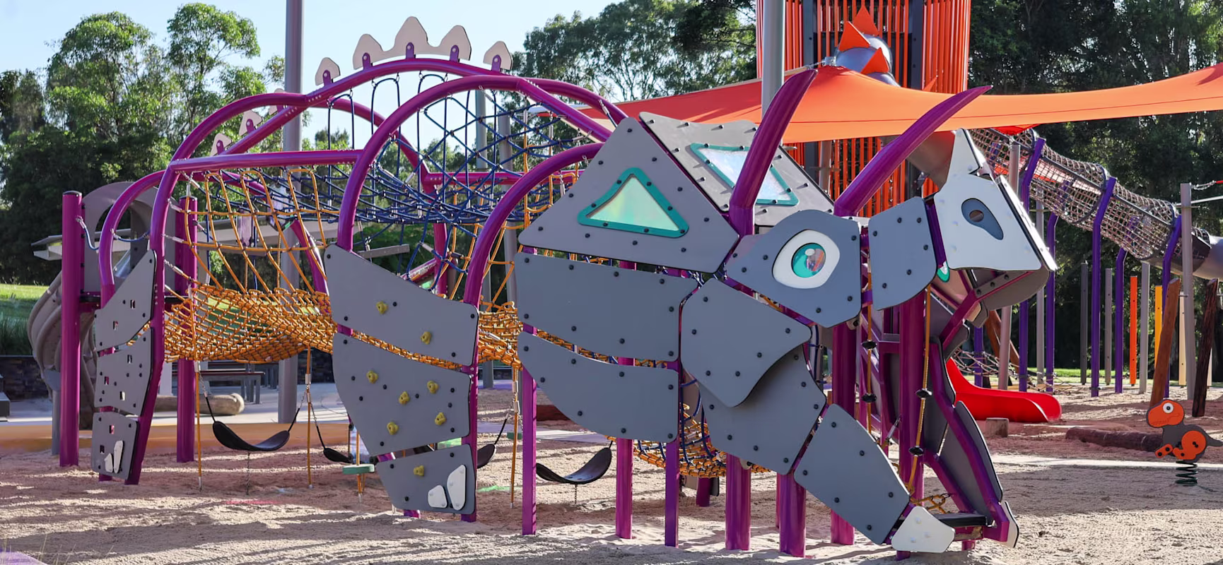 Colourful dinosaur-themed playground climbing structure with nets and slides under orange shade sails in a sandy play area.