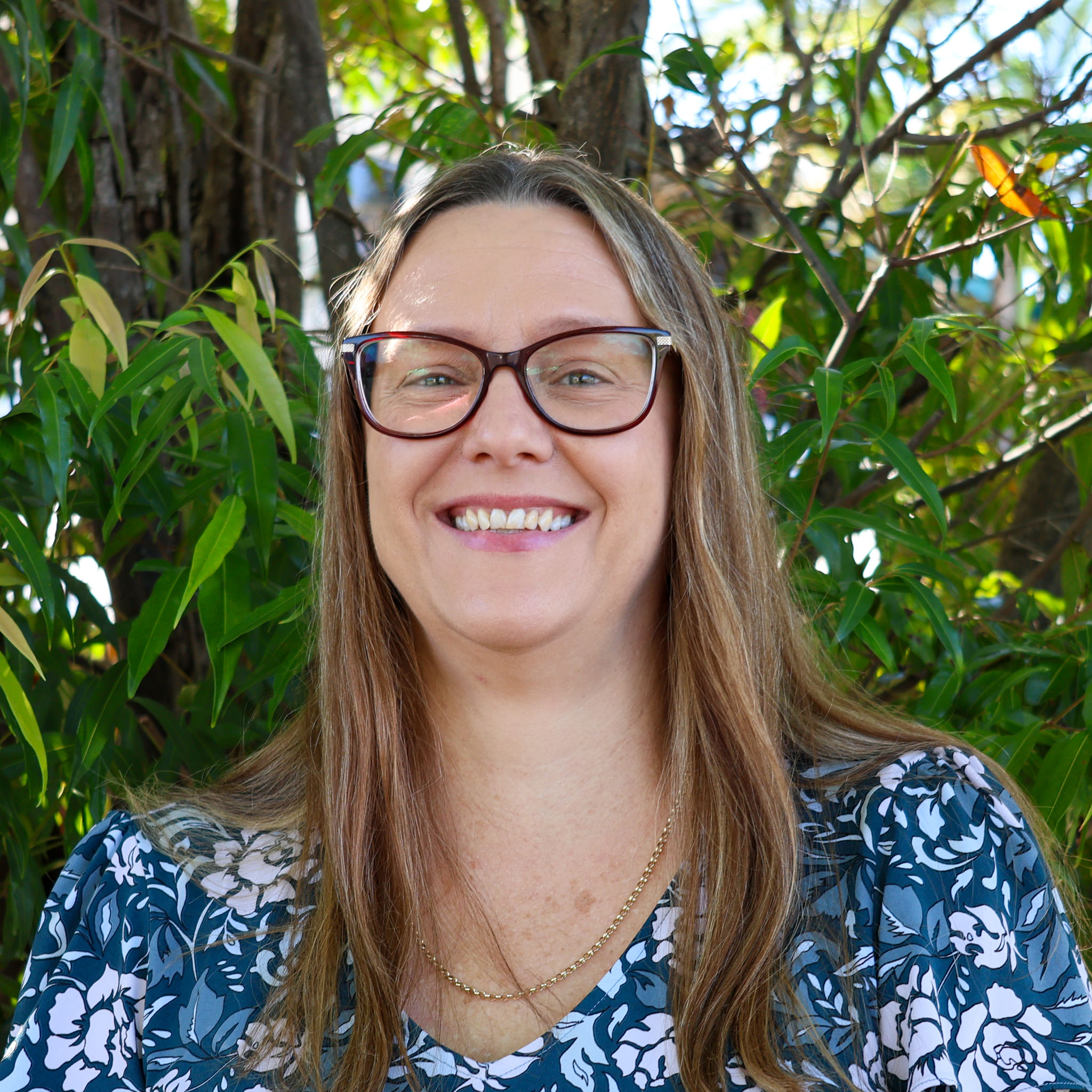 Person with blurred face, long light brown hair and floral top stands outdoors in front of green leafy trees.