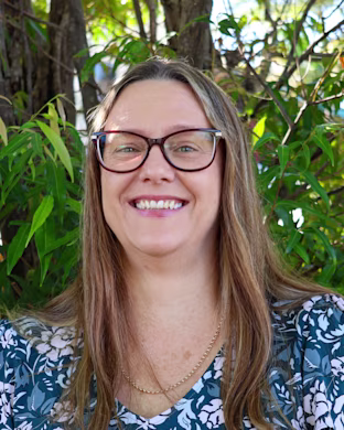 Person with blurred face, long light brown hair and floral top stands outdoors in front of green leafy trees.