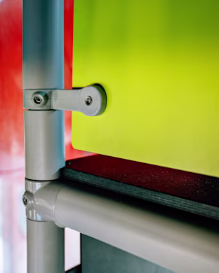 Close-up of grey metal pipes and brackets supporting a bright yellow panel, with red and dark surfaces in the background.