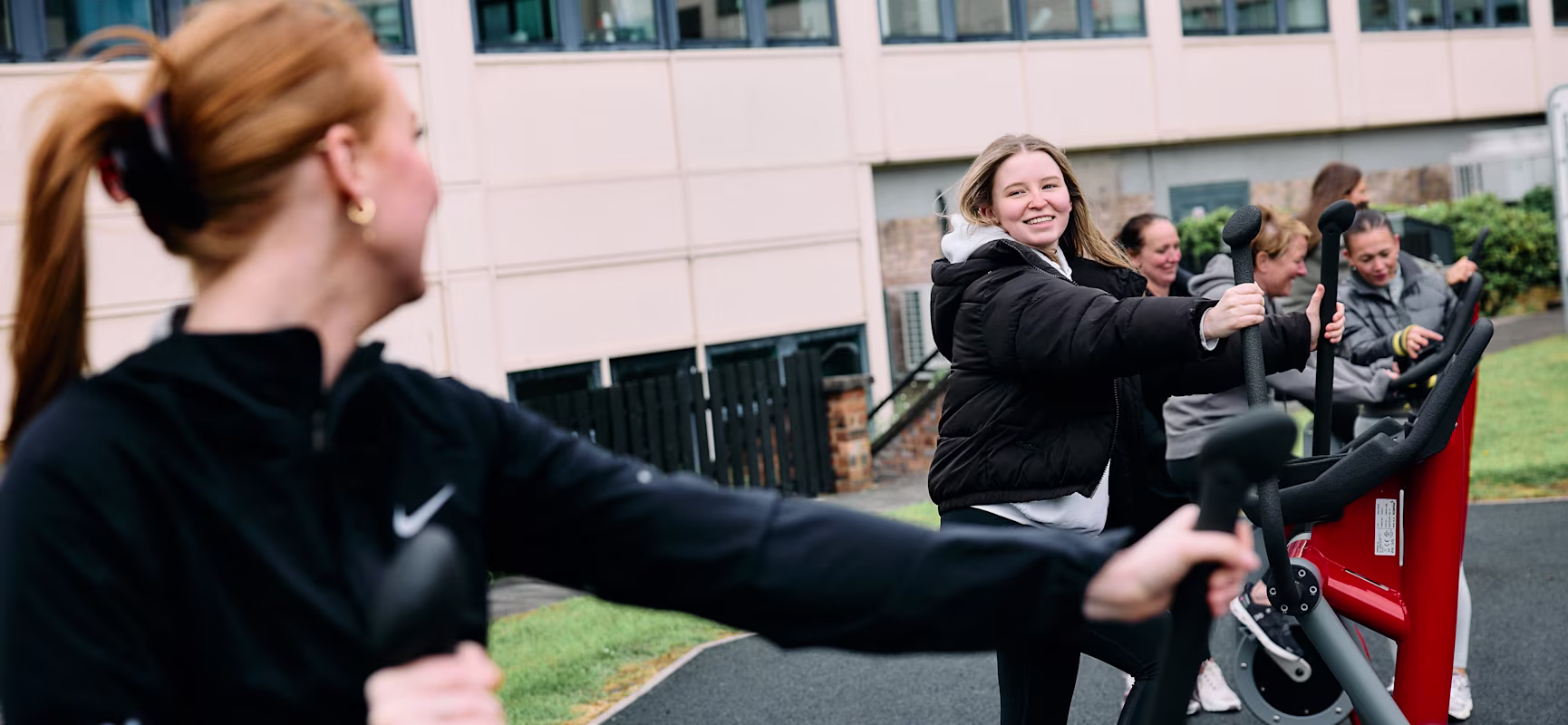 Group of people exercising on outdoor cardio machines beside a modern building, wearing jackets and sportswear on a cloudy day