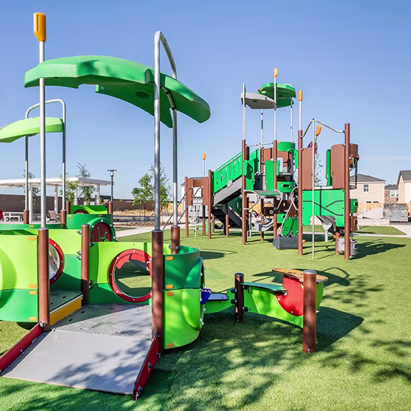 Woman helps two young children stand on a green spinning playground dish in an outdoor play area with modern equipment.