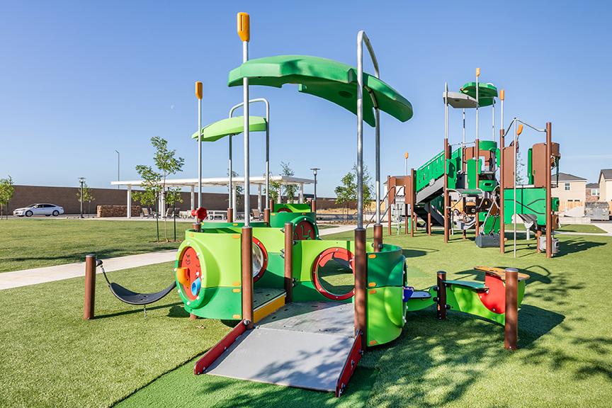 Woman helps two young children stand on a green spinning playground dish in an outdoor play area with modern equipment.