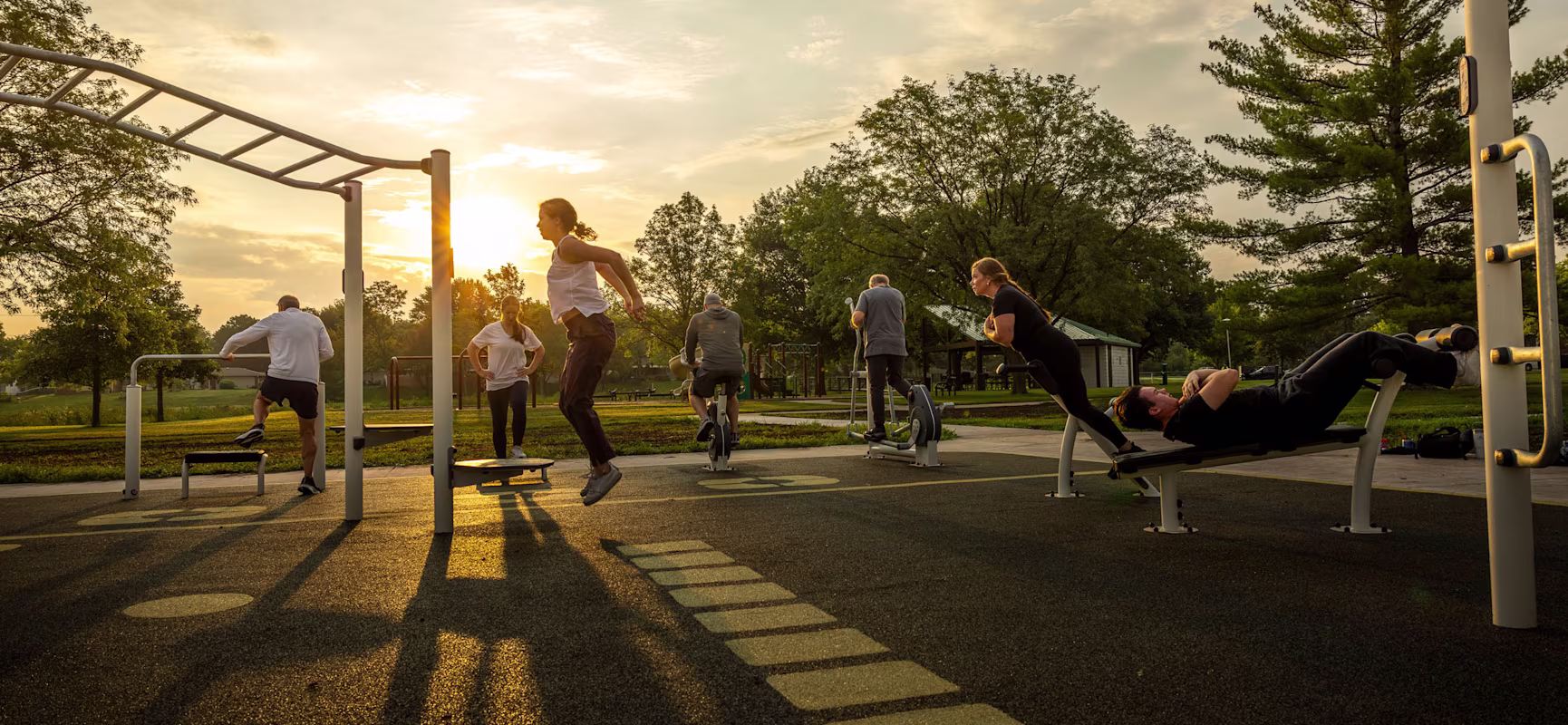 Outdoor fitness area with exercise machines and bars on a lawn, surrounded by trees and modern apartment buildings in bright daylight