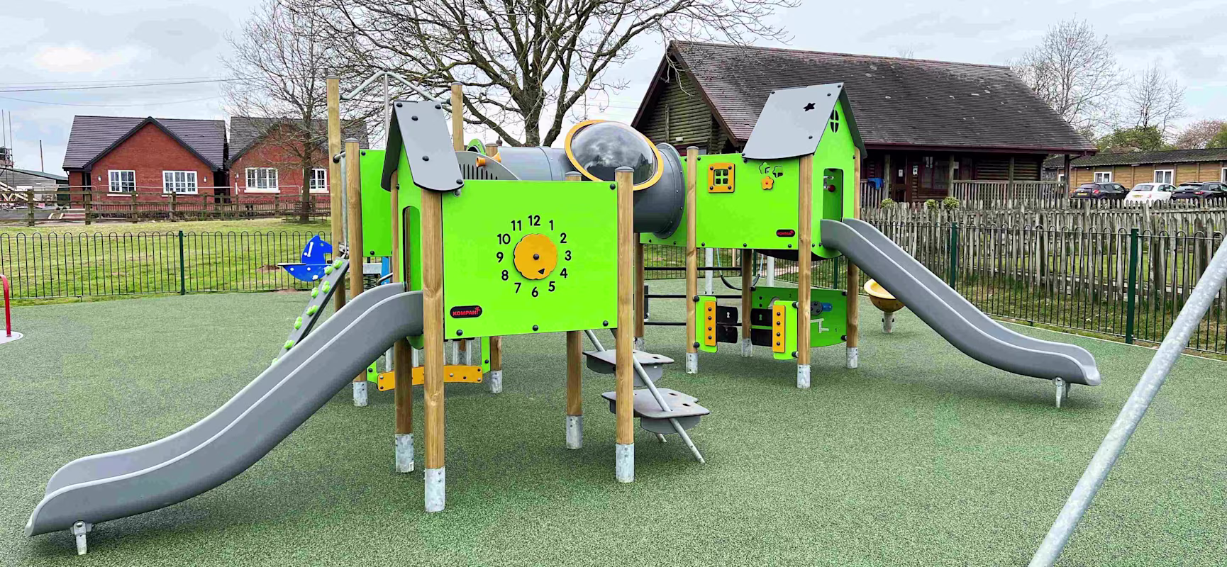 Empty playground with bright green climbing frame, two grey slides and tunnel, set on soft surface with houses and leafless trees behind