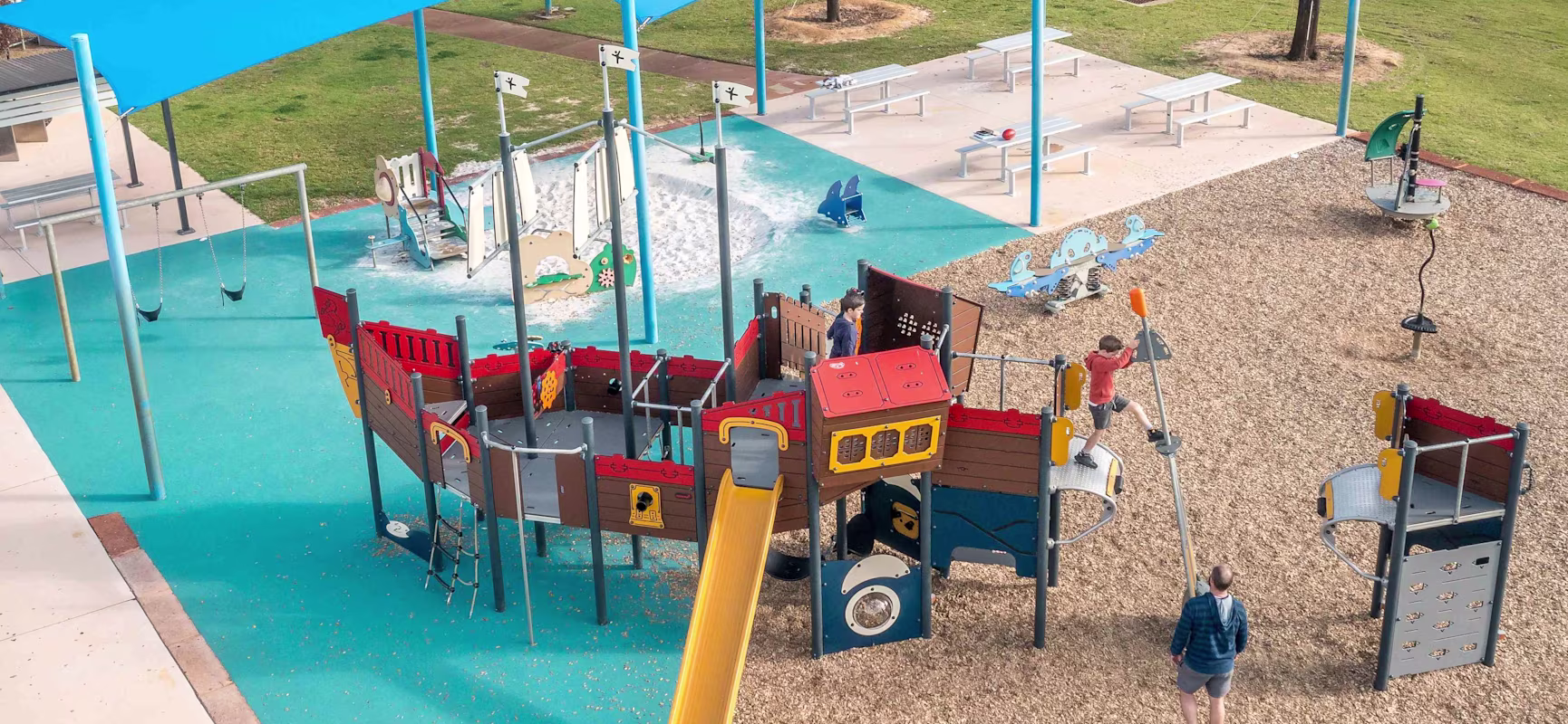 Aerial view of children playing on a colourful ship-themed playground under blue shade sails in a park.