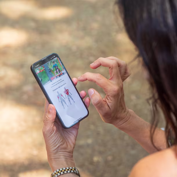 Woman with red nails uses smartphone outdoors, viewing a fitness app screen with exercise images and muscle impact diagram.