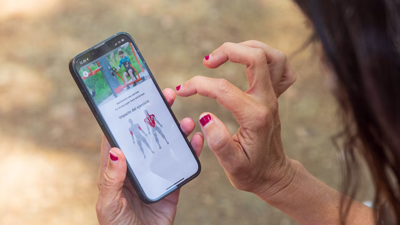 Woman with red nails uses smartphone outdoors, viewing a fitness app screen with exercise images and muscle impact diagram.
