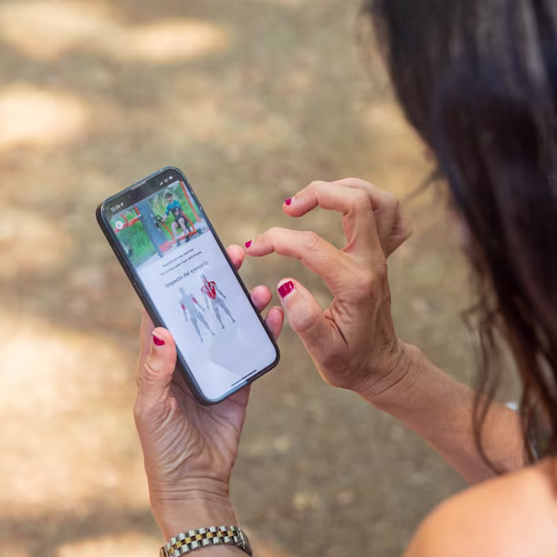 Woman with red nails uses smartphone outdoors, viewing a fitness app screen with exercise images and muscle impact diagram.