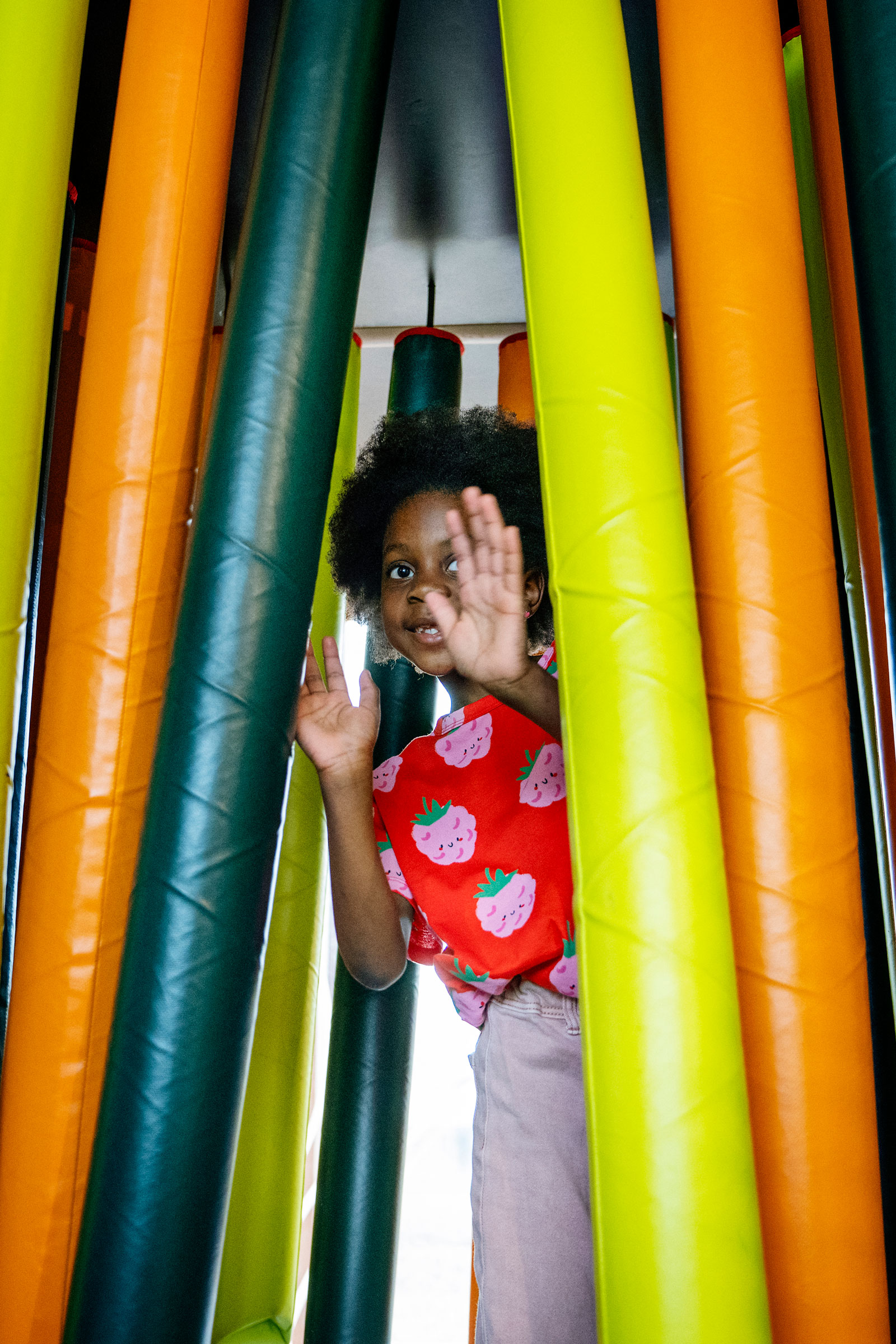 Child in strawberry shirt playing among tall colorful foam tubes, peeking through and raising hands inside an indoor play area