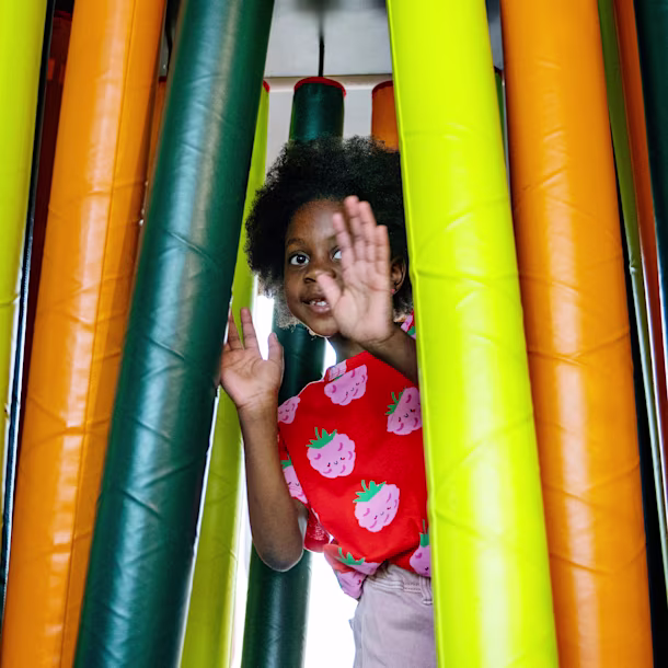 Child in strawberry shirt playing among tall colorful foam tubes, peeking through and raising hands inside an indoor play area
