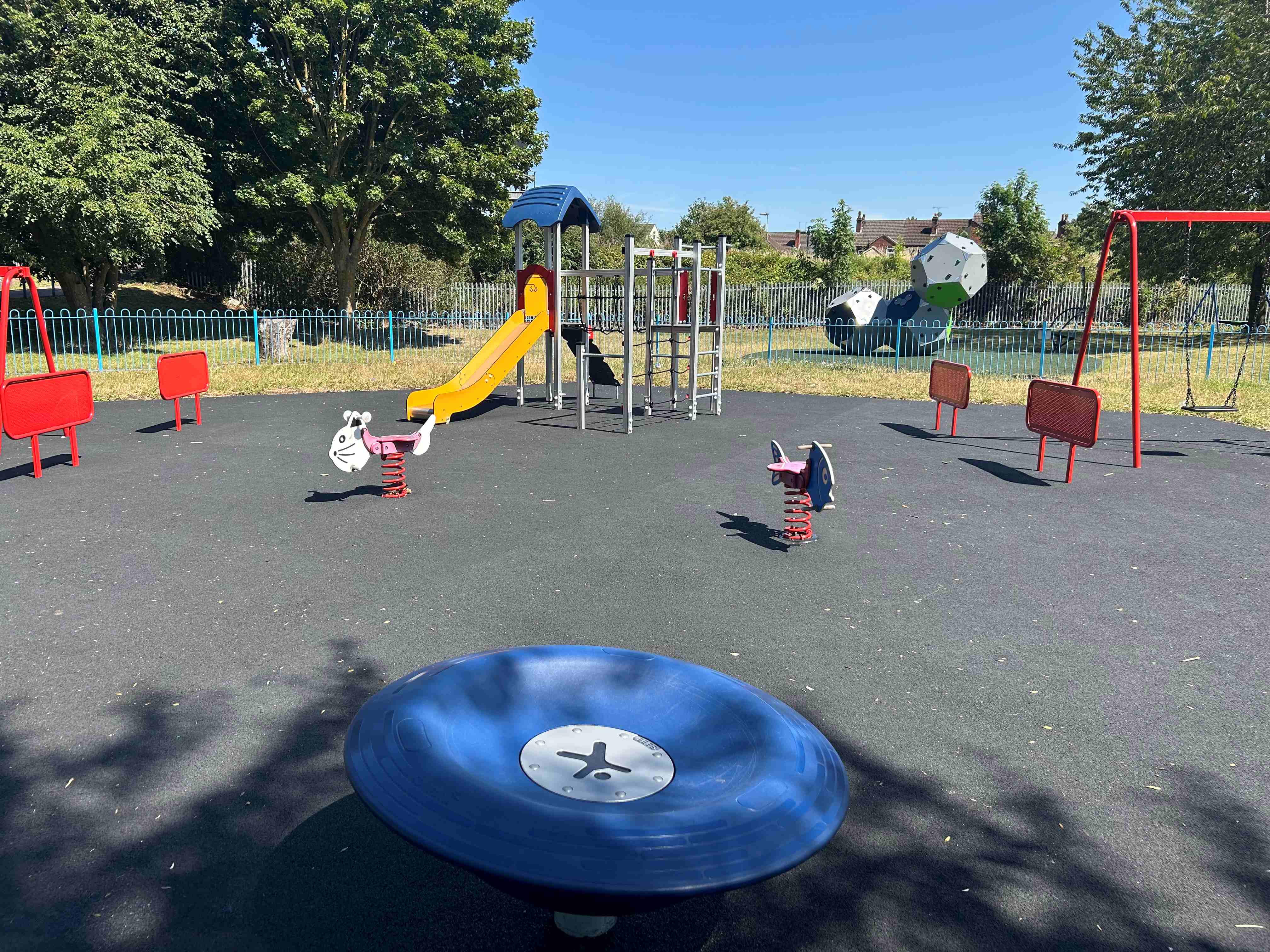 Sunny playground with slide, swings, round blue spinner and spring riders on rubber surface, trees and houses in the background
