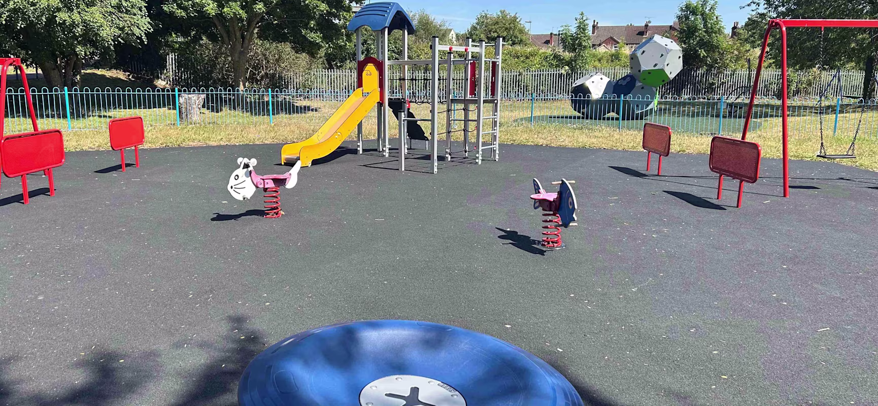 Sunny playground with slide, swings, round blue spinner and spring riders on rubber surface, trees and houses in the background
