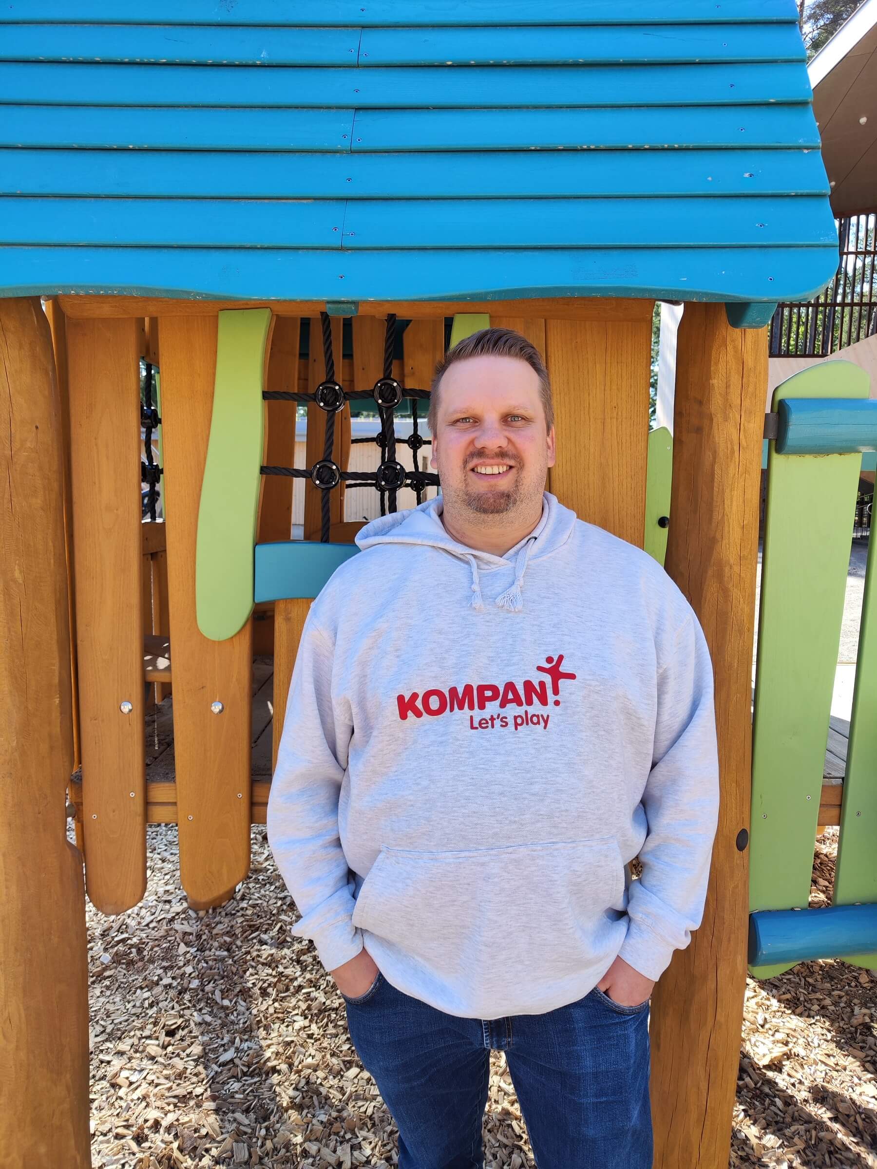 Person in grey “KOMPAN Let’s play” hoodie stands in colourful wooden playground structure with blue roof and bark ground.