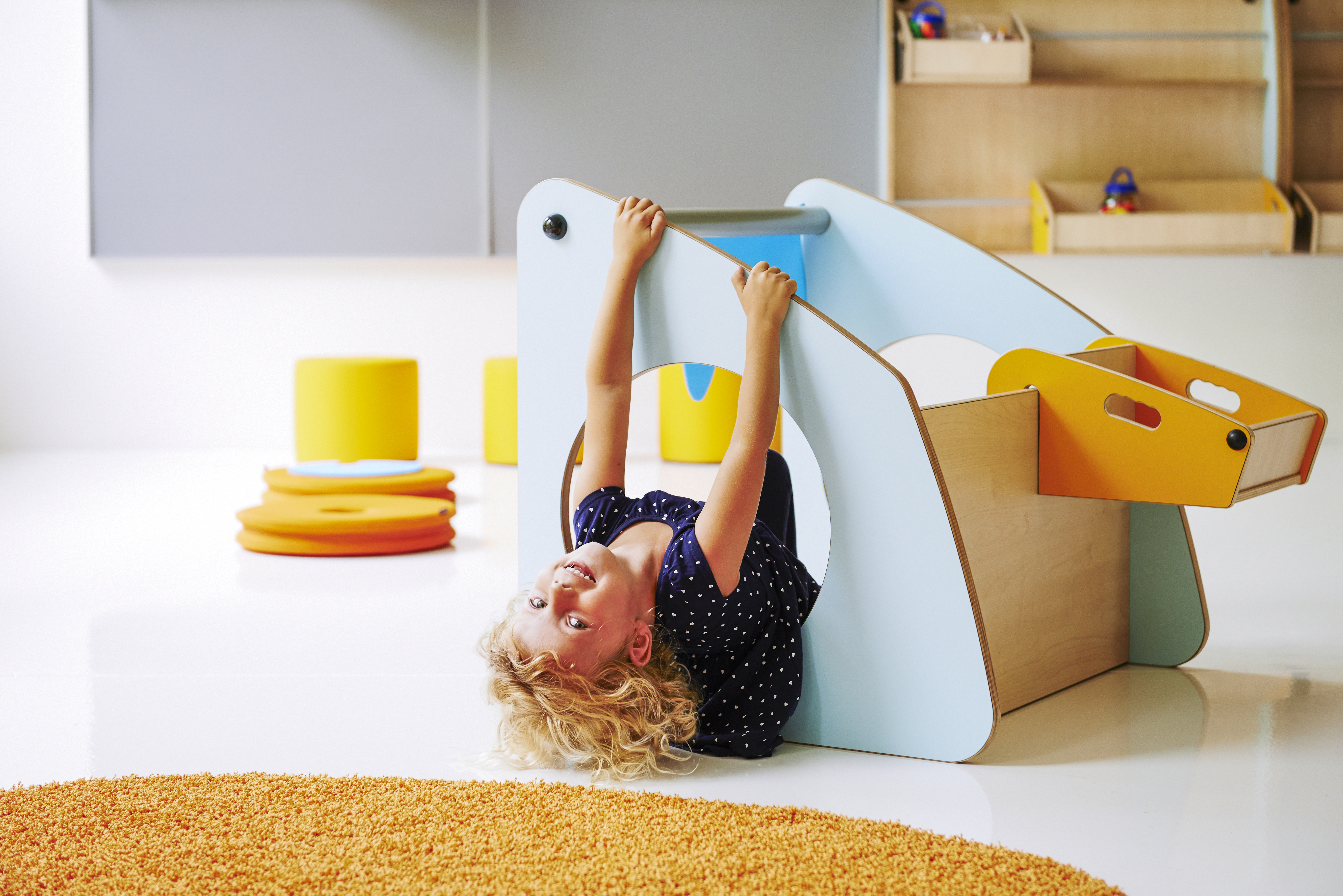 Child hanging upside down through a hole in a blue play structure in a bright playroom with yellow and orange soft blocks.