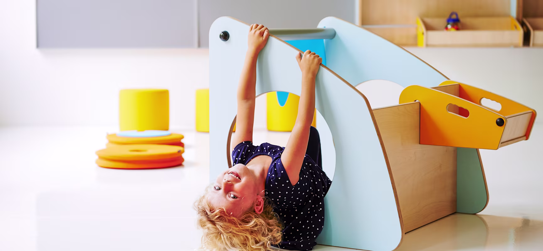 Child hanging upside down through a hole in a blue play structure in a bright playroom with yellow and orange soft blocks.