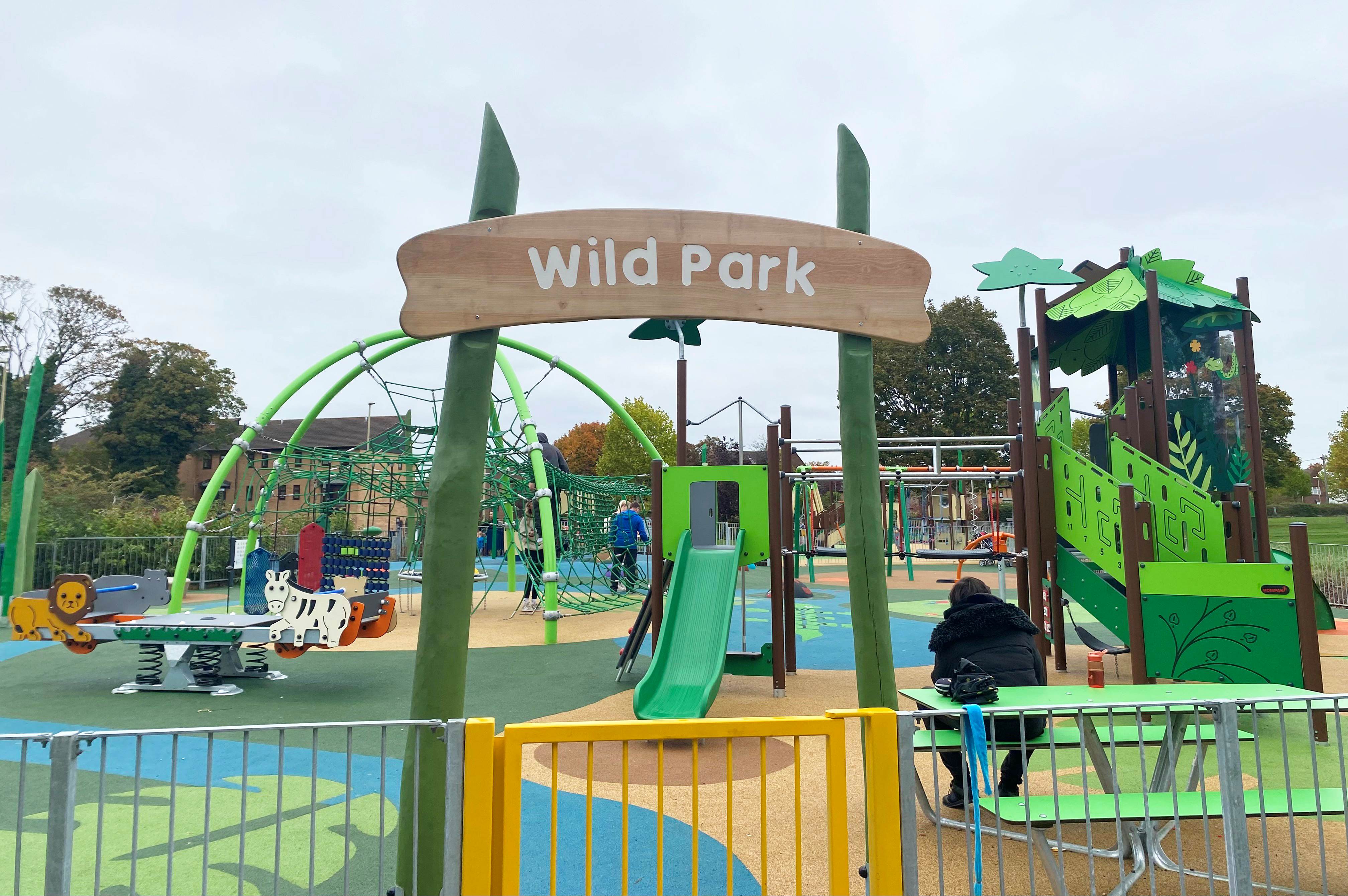 Colourful “Wild Park” playground with slides, climbing nets, animal-themed ride-ons and a seated adult near a green picnic table