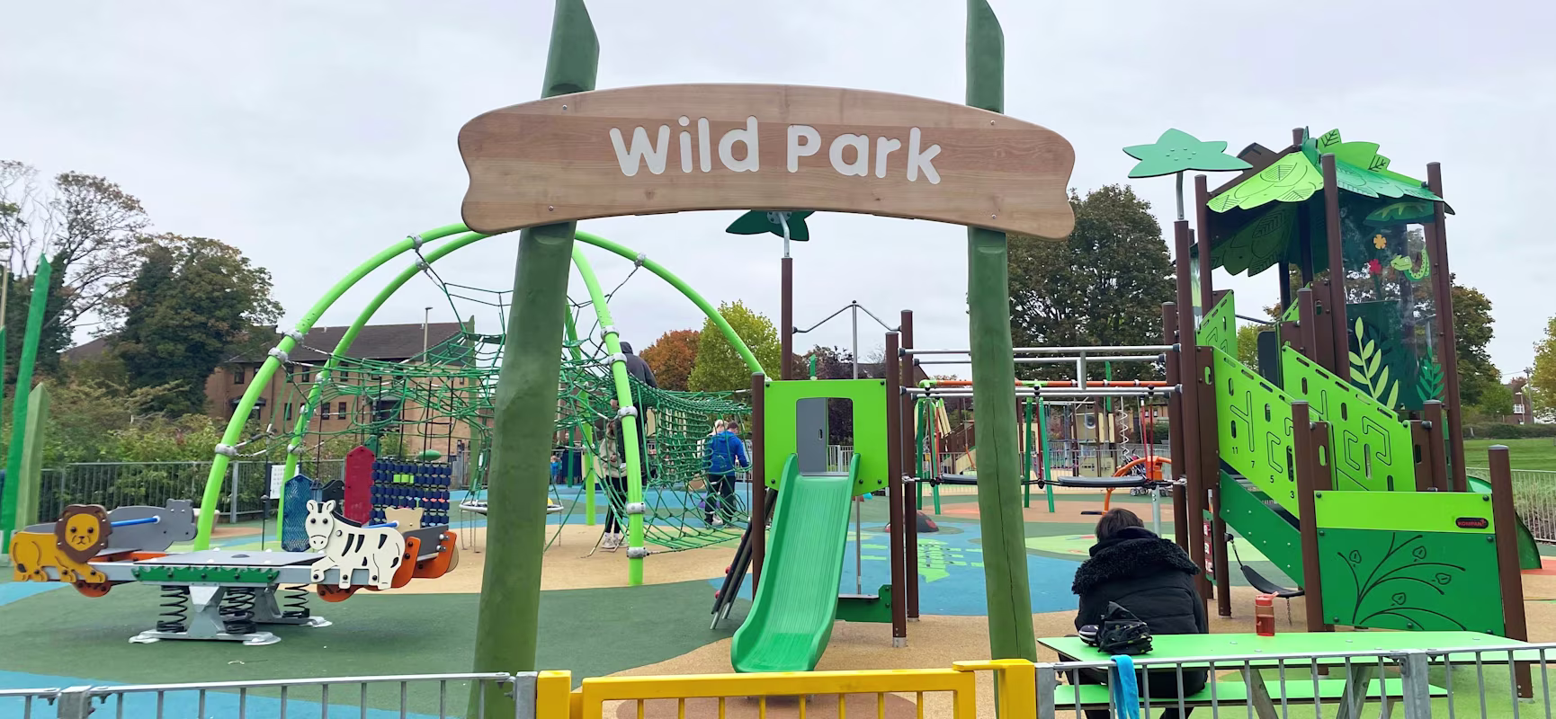 Colourful “Wild Park” playground with slides, climbing nets, animal-themed ride-ons and a seated adult near a green picnic table