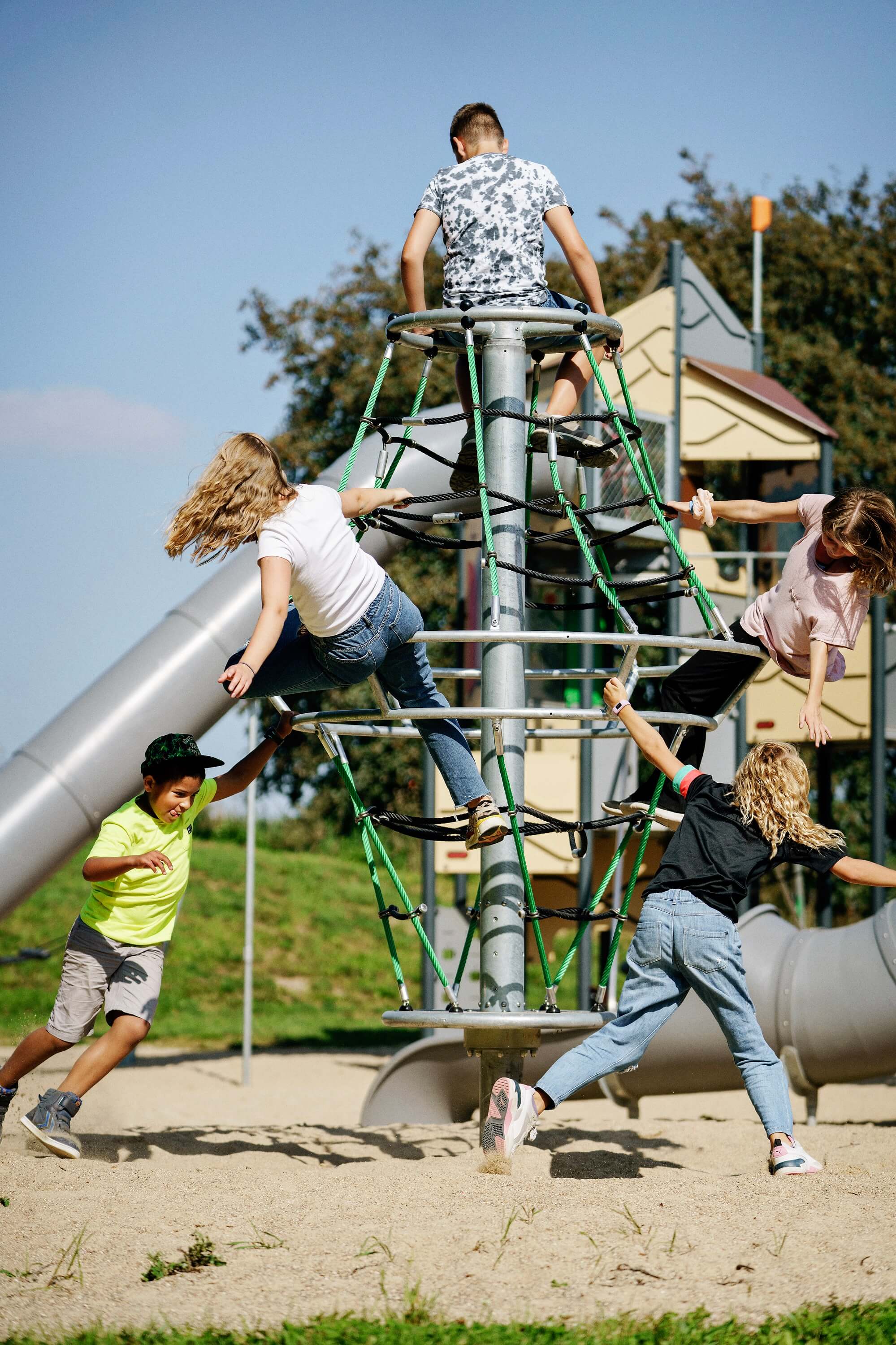 Children climb and play on a tall rope structure in a sunny playground with slides and sand underneath