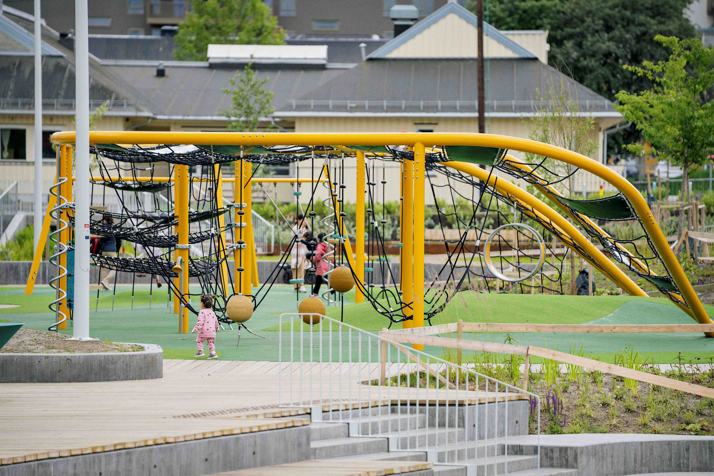 Children climb and play on a tall rope structure in a sunny playground with slides and sand underneath