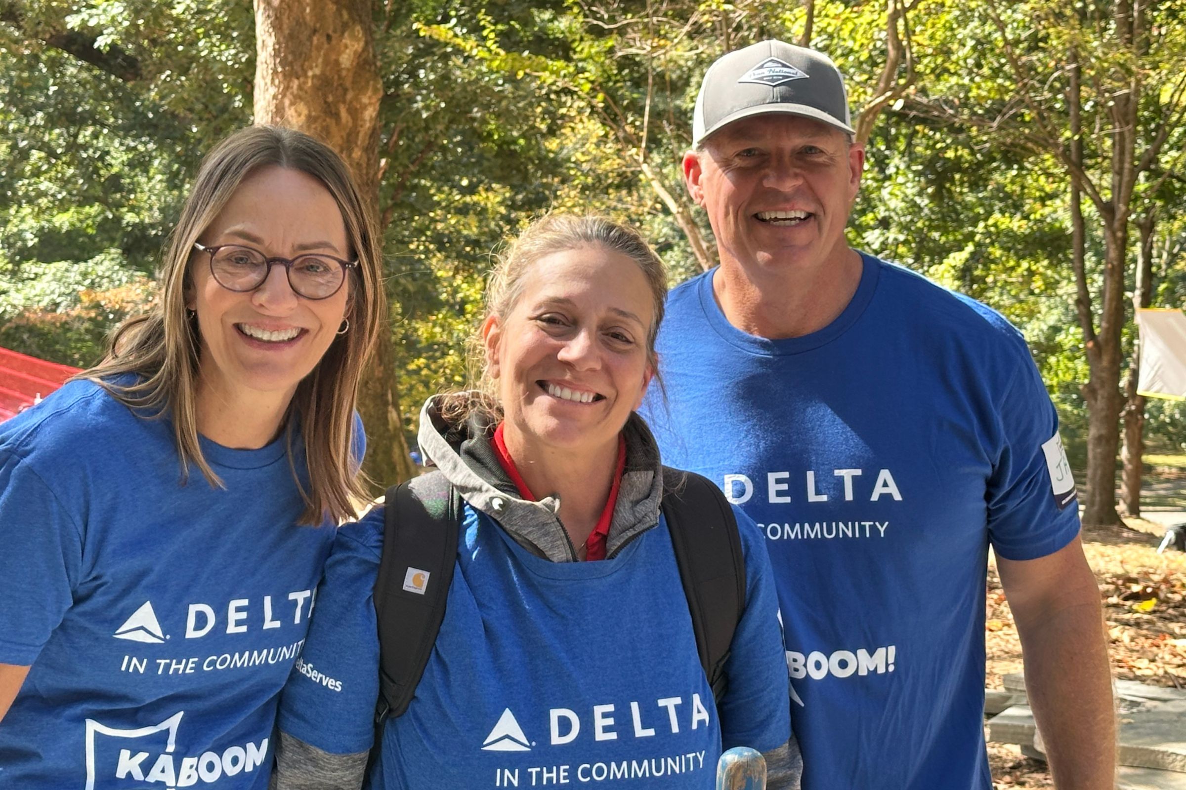 Three volunteers outdoors wearing matching blue “Delta in the Community” shirts, standing close together among trees in daylight.