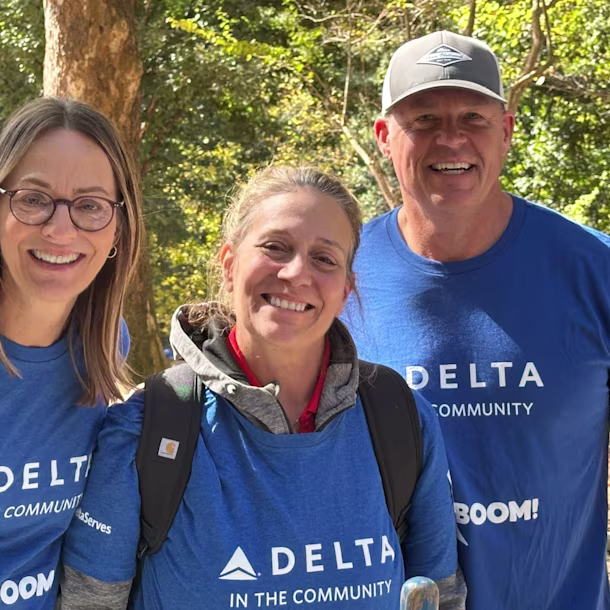 Three volunteers outdoors wearing matching blue “Delta in the Community” shirts, standing close together among trees in daylight.