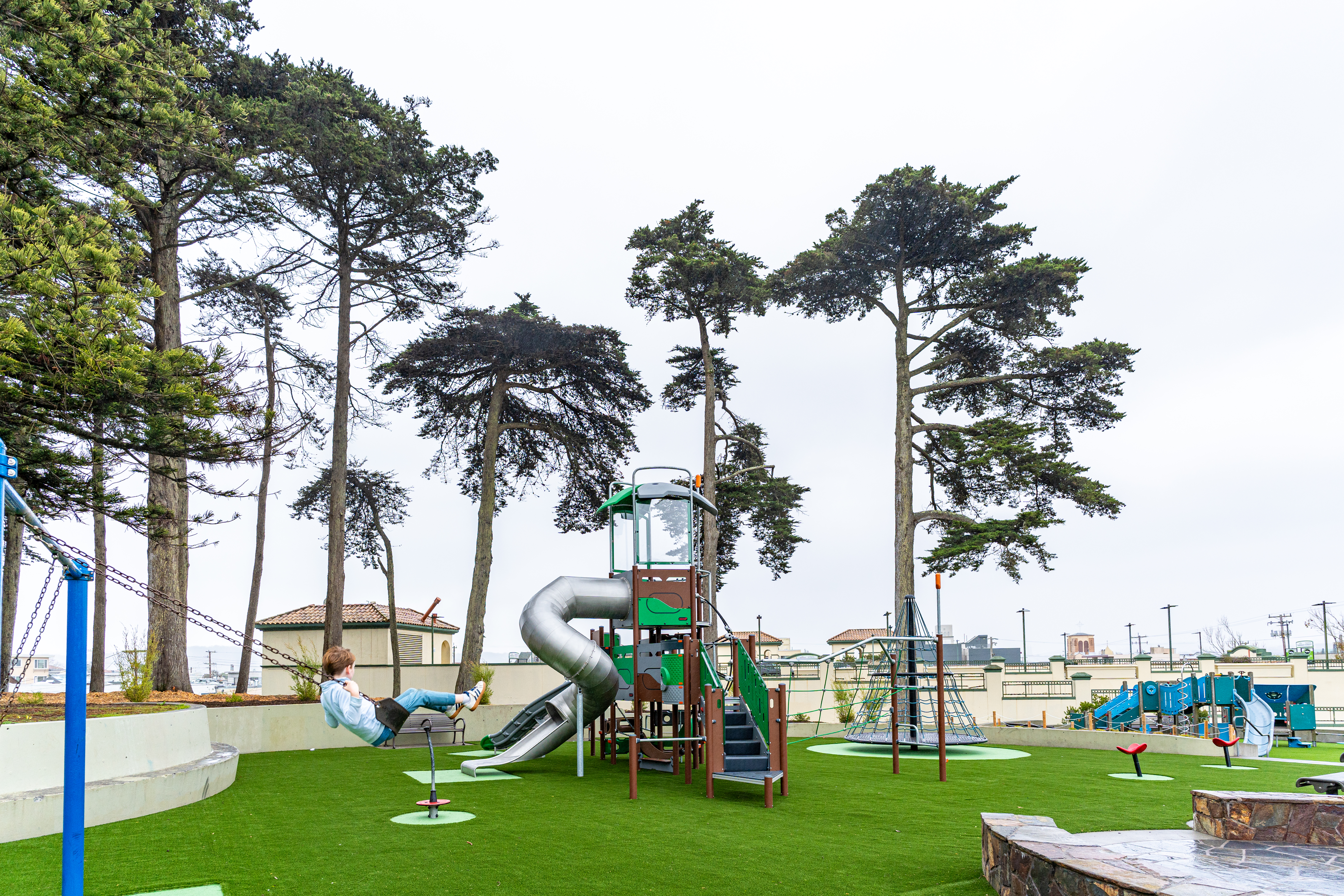 Wide shot of Lincoln Playground in San Francisco, California, featuring a child swinging and swaying trees in the background on an overcast day