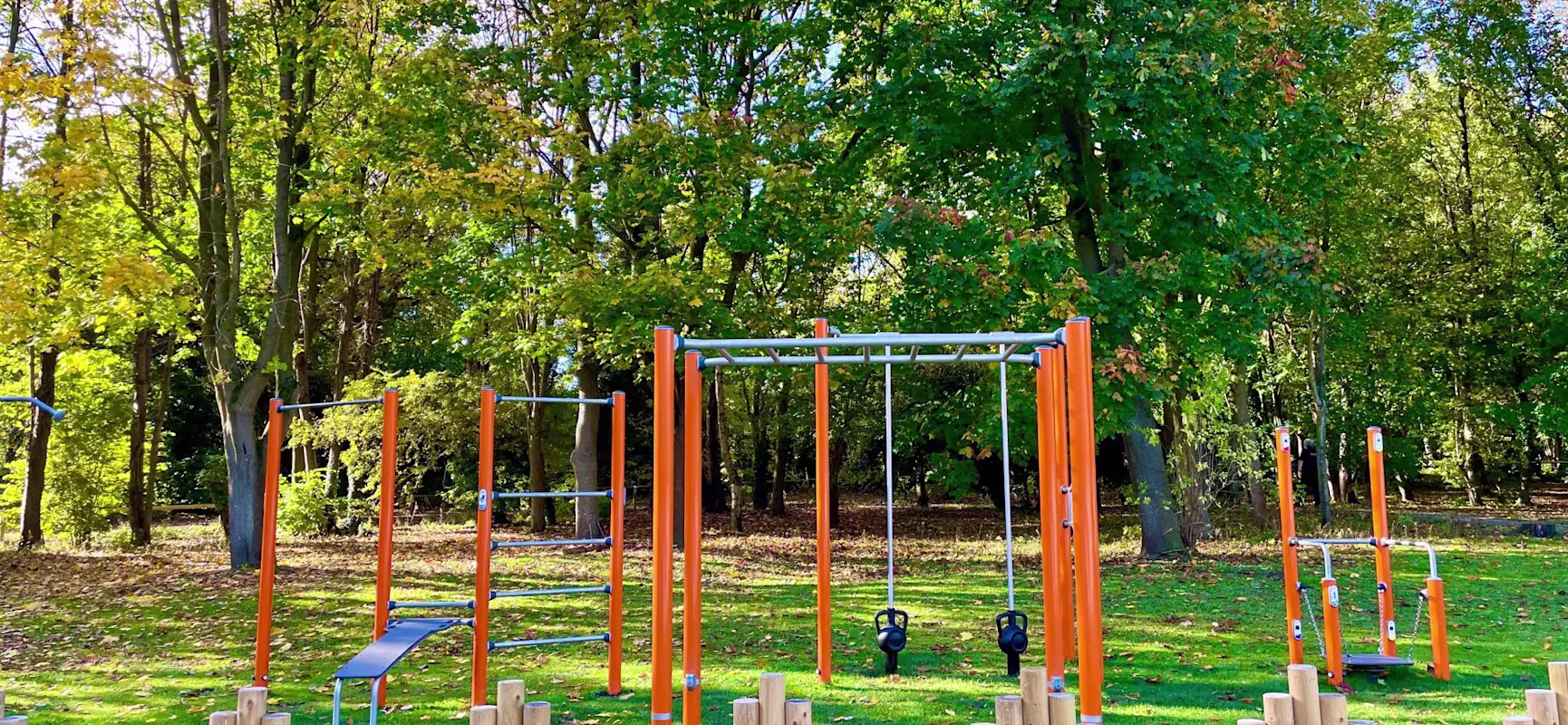 Outdoor fitness park with bright orange workout bars and kettlebells on green grass, surrounded by tall trees and a low wooden fence