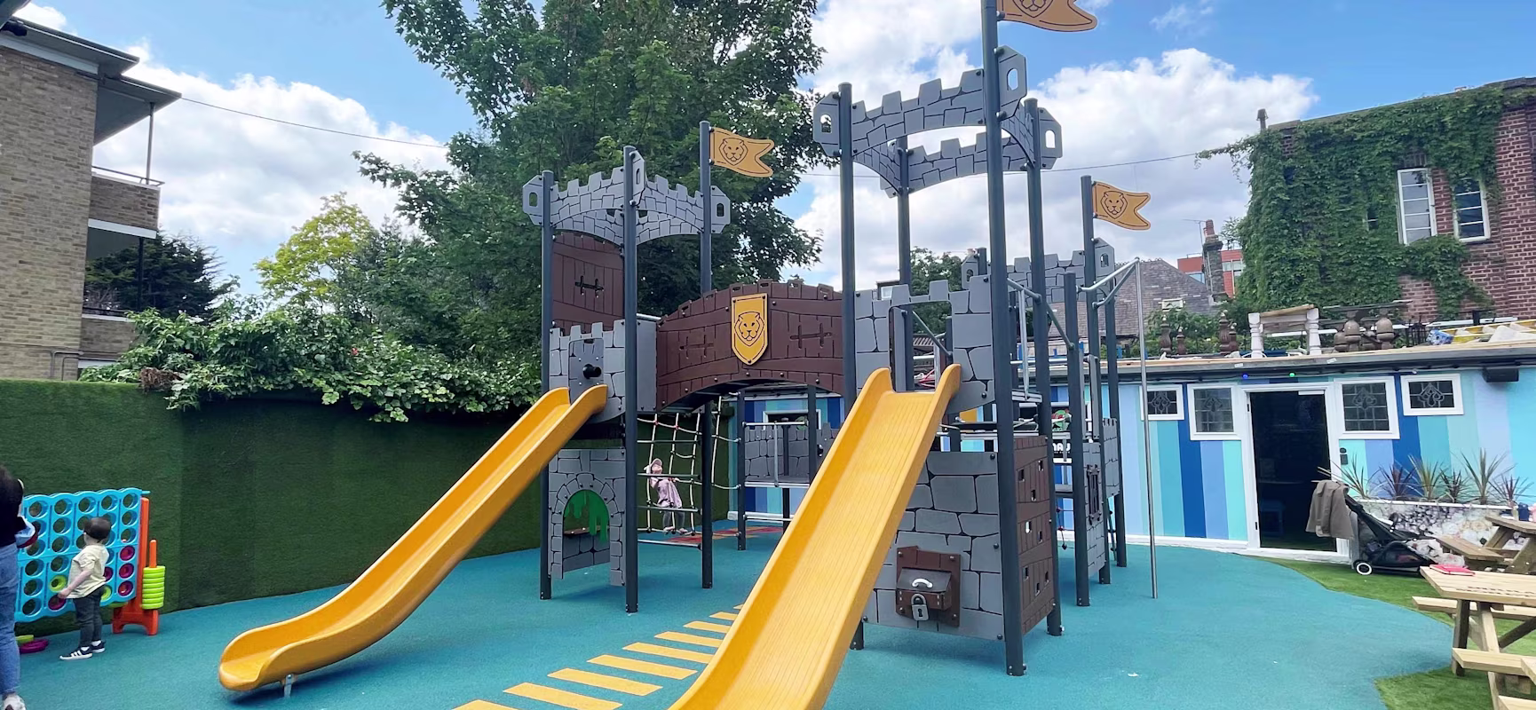 Castle-themed playground with two bright yellow slides on blue rubber flooring, children playing nearby and colourful buildings behind.