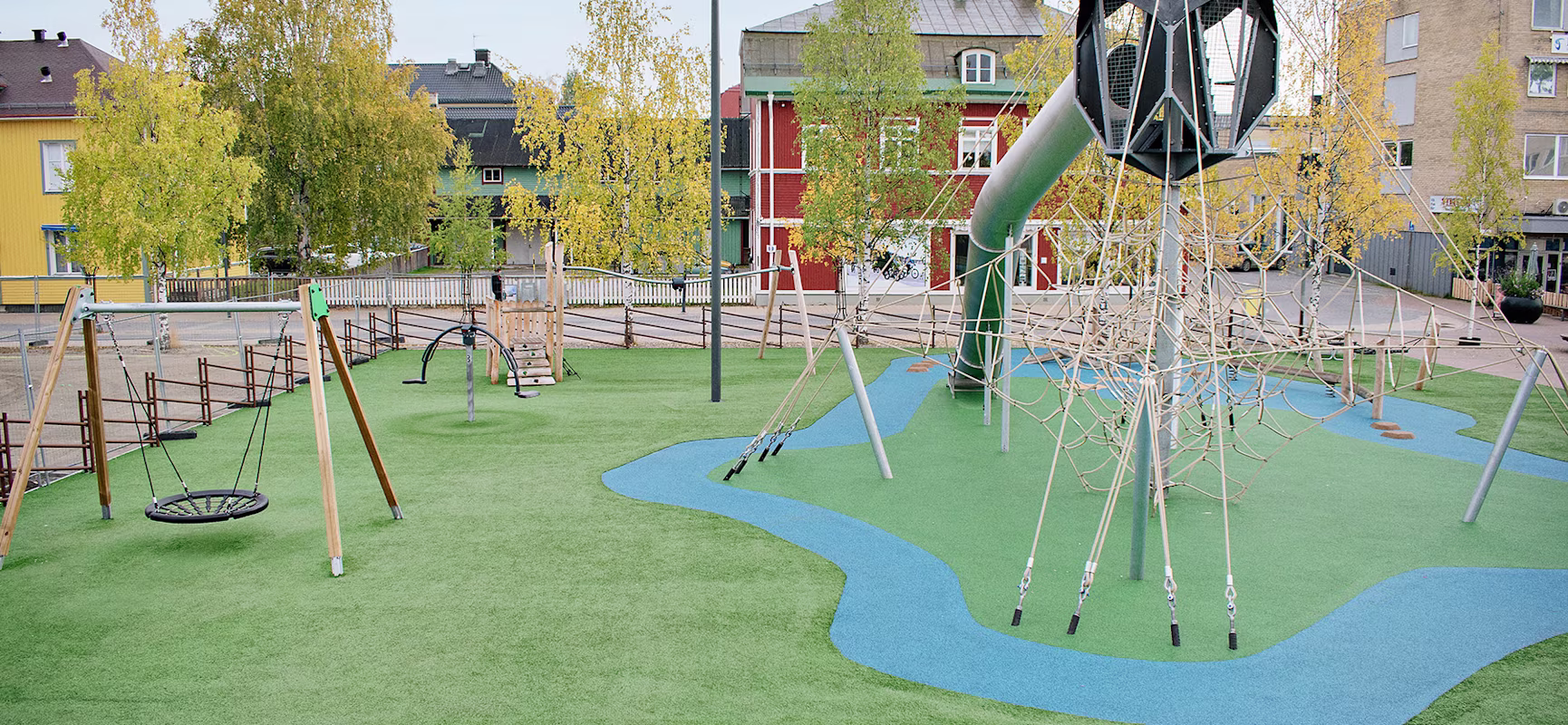 Modern outdoor playground with rope climbing tower, slide, swings and blue rubber paths on green turf, surrounded by trees and houses