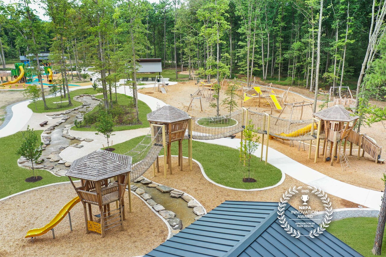 Award-winning forest playground with wooden towers, rope bridges, yellow slides, winding stream, and walking paths among tall trees.