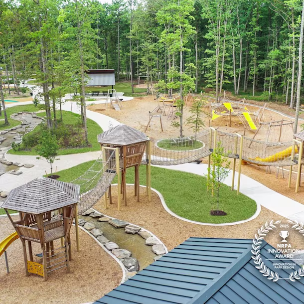 Award-winning forest playground with wooden towers, rope bridges, yellow slides, winding stream, and walking paths among tall trees.