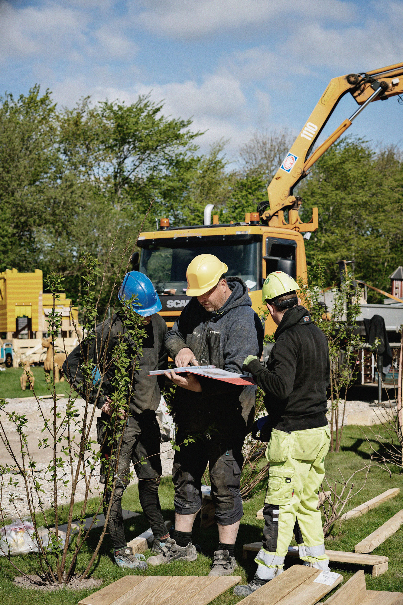 Three construction workers in helmets review plans on a clipboard at a grassy worksite, with a yellow truck and excavator behind them.