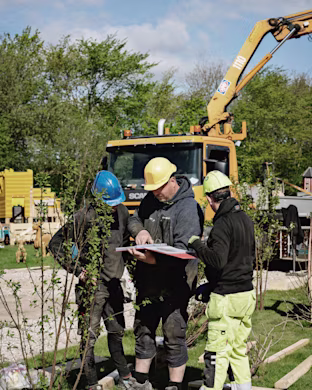 Three construction workers in helmets review plans on a clipboard at a grassy worksite, with a yellow truck and excavator behind them.