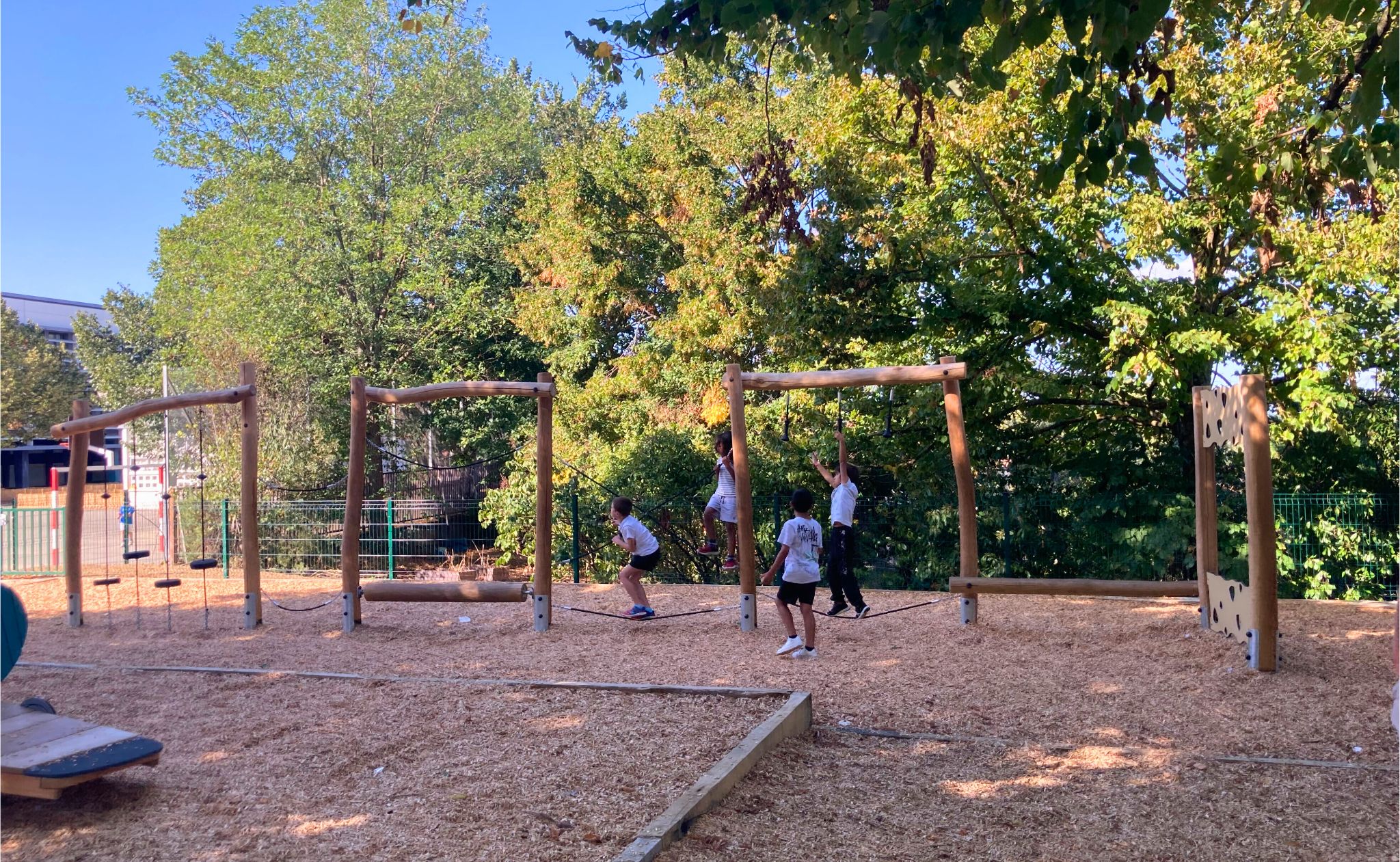 Children playing on a wooden climbing and balance structure in a sunny playground surrounded by green trees.