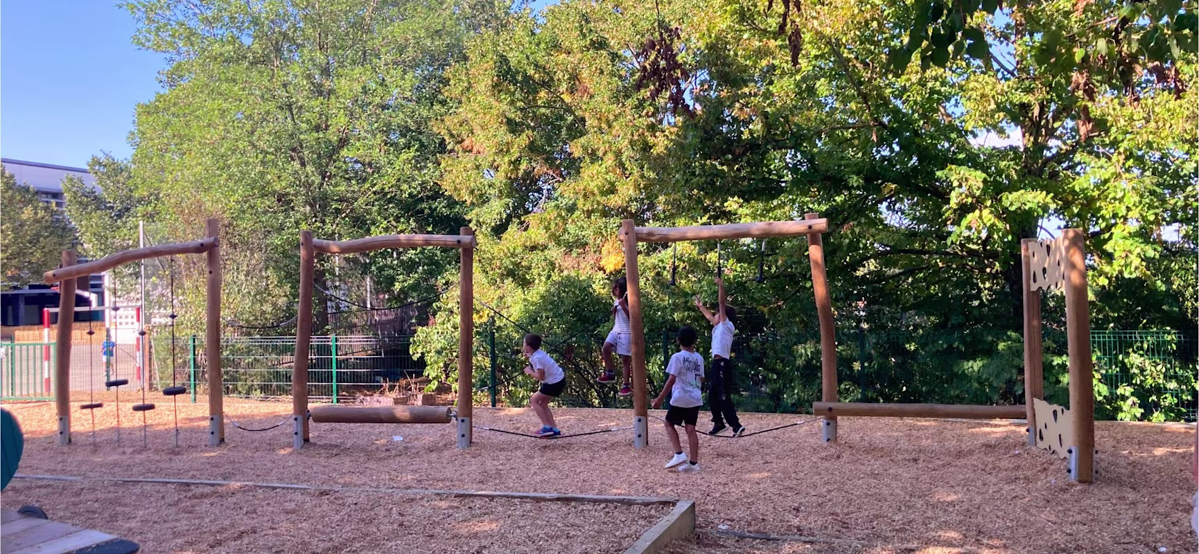 Children playing on a wooden climbing and balance structure in a sunny playground surrounded by green trees.