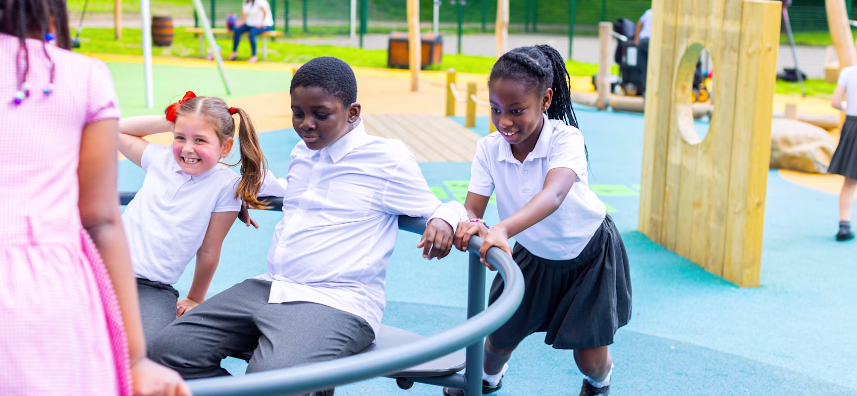 Schoolchildren in uniforms play on a spinning roundabout in a colourful outdoor playground with climbing frames in the background.
