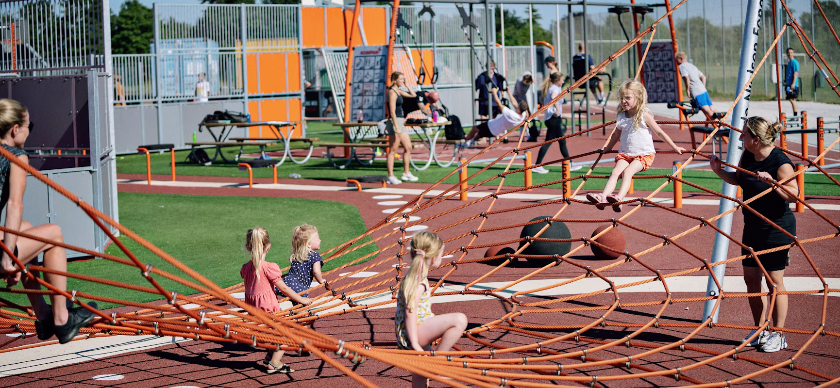 Children play on a large wooden playground with rope bridge, slides and climbing bars, surrounded by trees and woodchip ground.