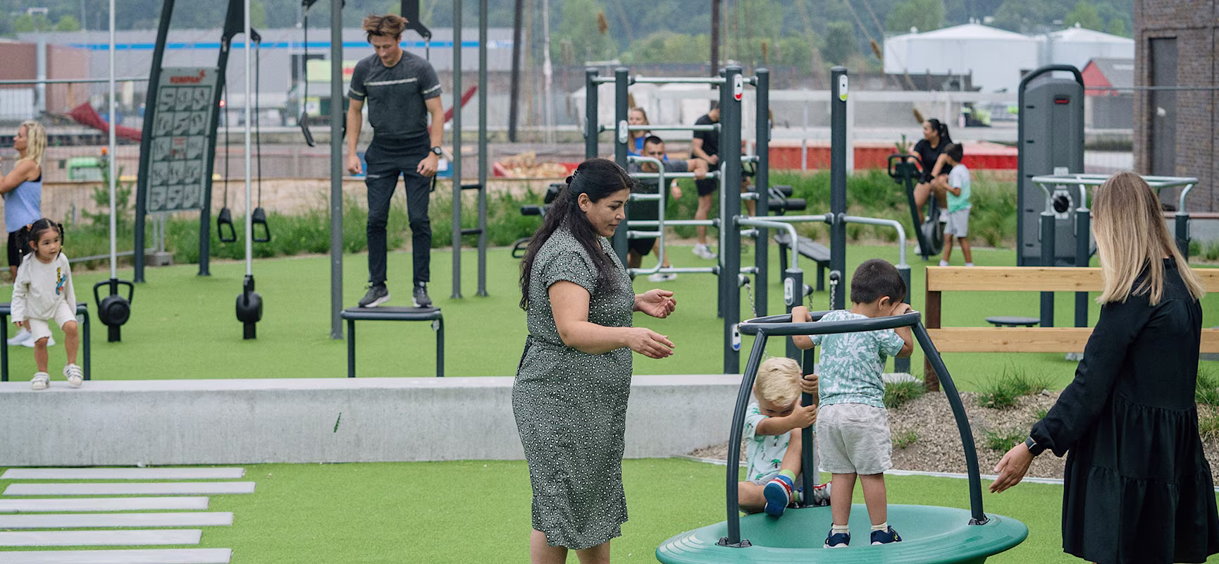 Adults and children play on outdoor fitness and playground equipment in a green park, with two boys on a round spinner in the foreground