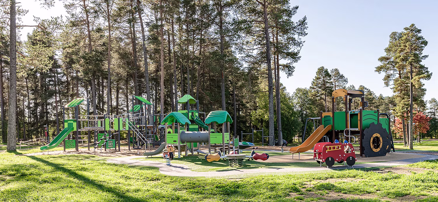 Sunny forest playground with green slides, climbing frames and a tractor-themed play structure on bright grass.