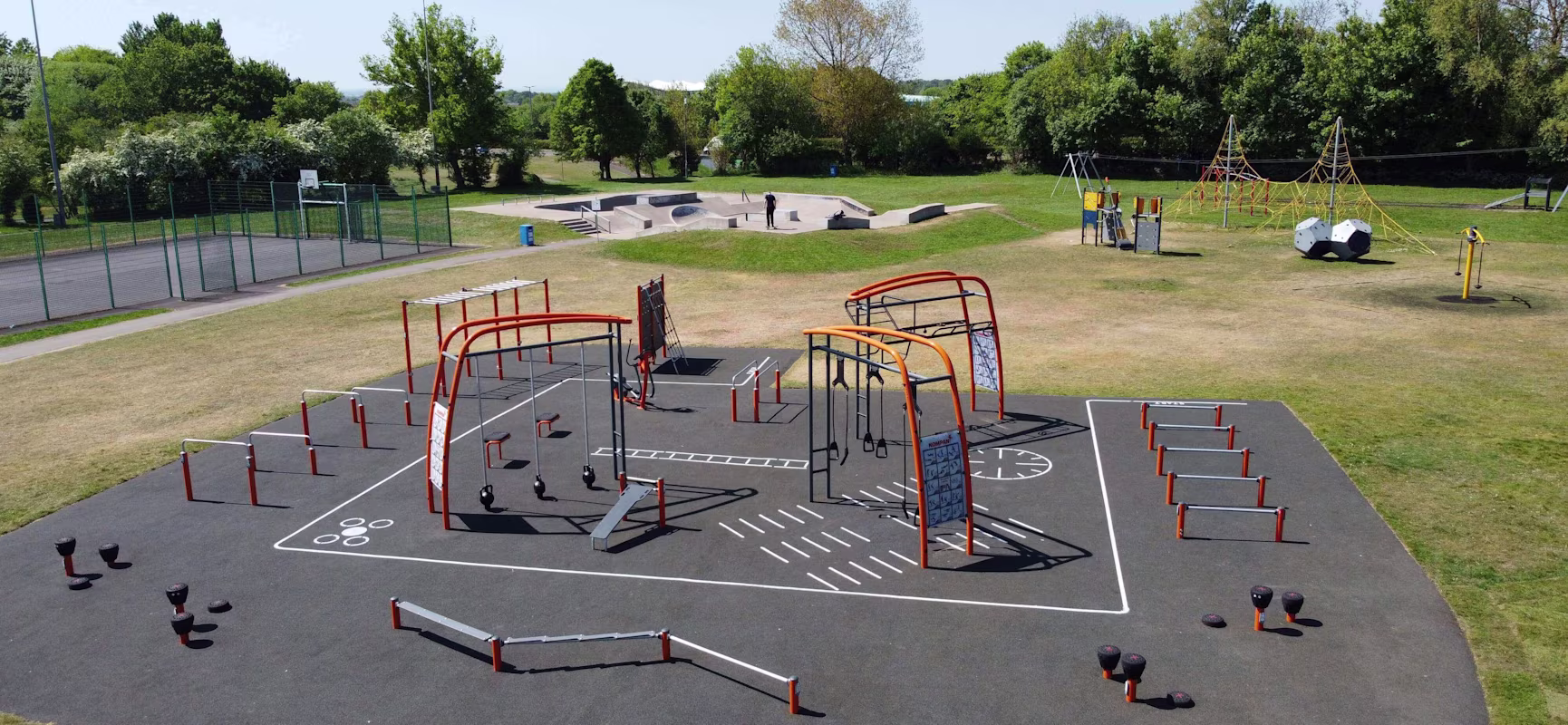 Large outdoor playground with orange and black climbing frames, swings, and balance equipment on rubber surface, surrounded by grass and trees