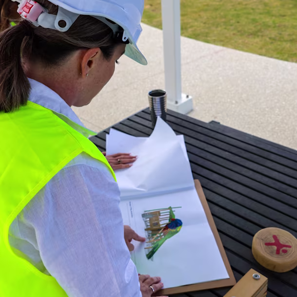 Person in a Kompan safety vest and hard hat reviews a colorful playground design on paper at an outdoor table.