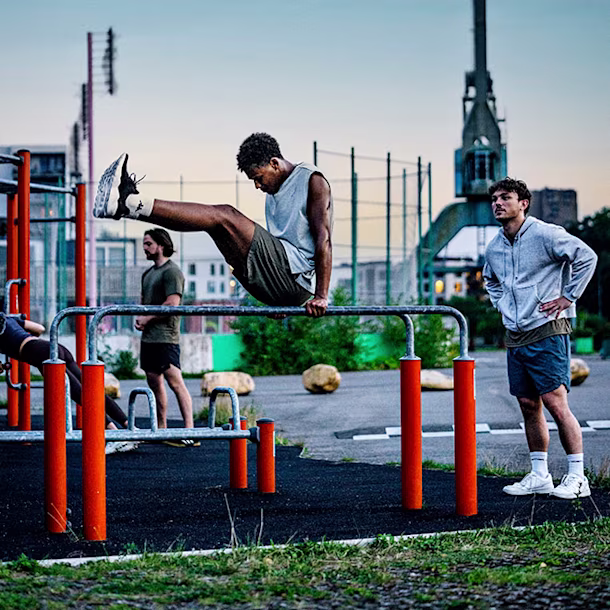 People exercise at an outdoor gym; two hang from monkey bars while others work out and stand nearby on a grey rubber surface.