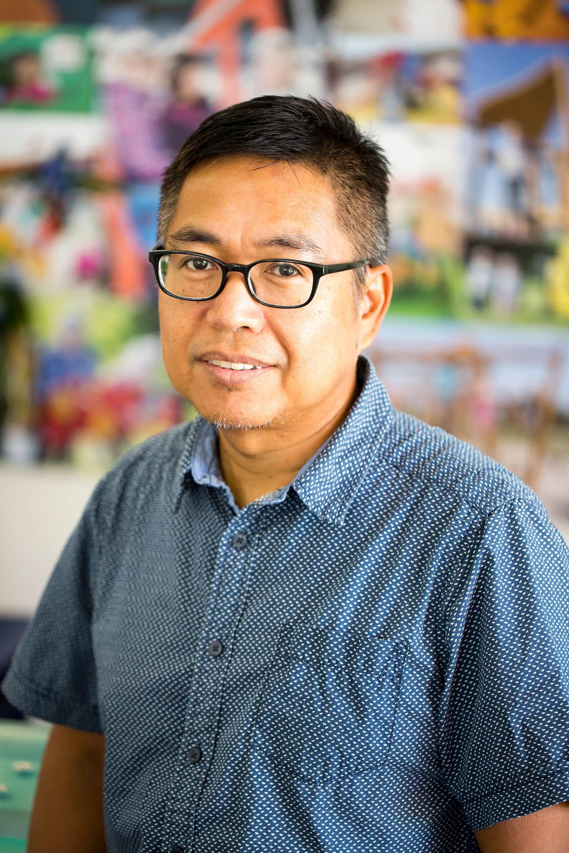Person in a blue patterned short-sleeve shirt, face blurred, standing indoors before a colourful, out-of-focus photo collage backdrop.