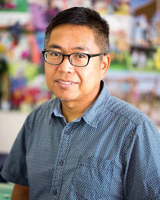 Person in a blue patterned short-sleeve shirt, face blurred, standing indoors before a colourful, out-of-focus photo collage backdrop.