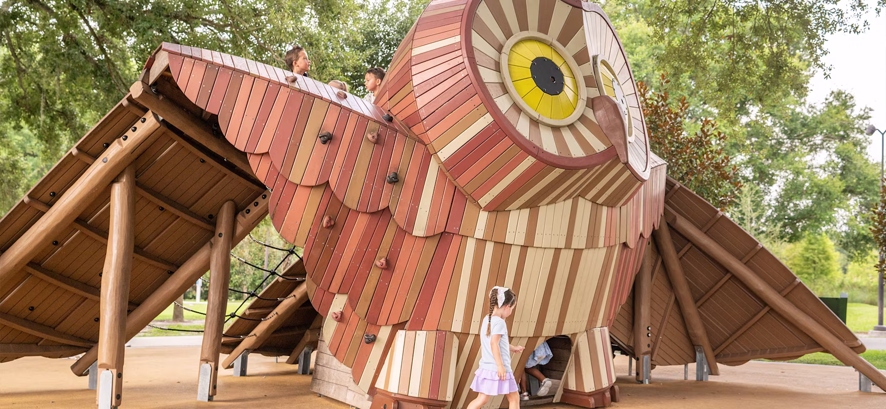 Children play on and inside a large wooden owl-shaped playground structure under leafy trees in a park.