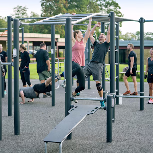 People exercise at an outdoor gym; two hang from monkey bars while others work out and stand nearby on a grey rubber surface.