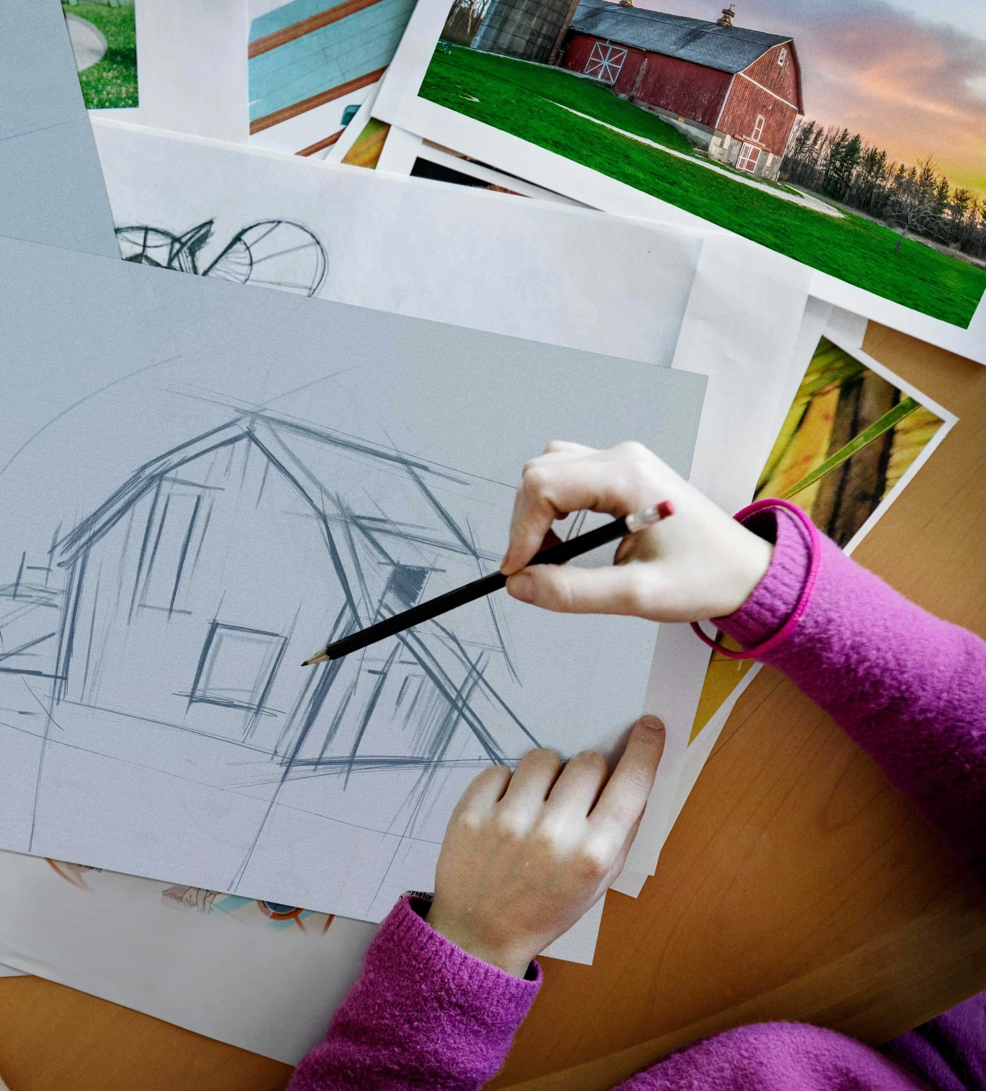 Overhead shot of a girl sketching a piece of playground equipment inspired by a house. She is sitting at a table. Reference images are spread across the table. Reference images can be a helpful playground planning tool.