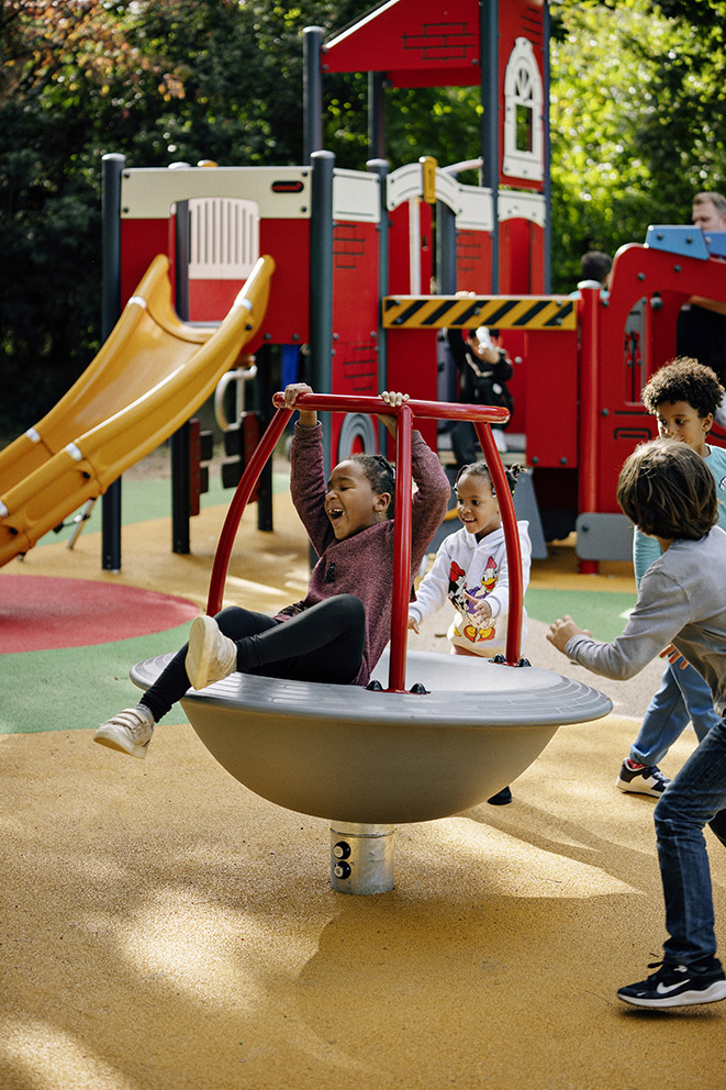 Children play on a spinning bowl ride in a colourful playground with a large red climbing structure and yellow slide in the background.